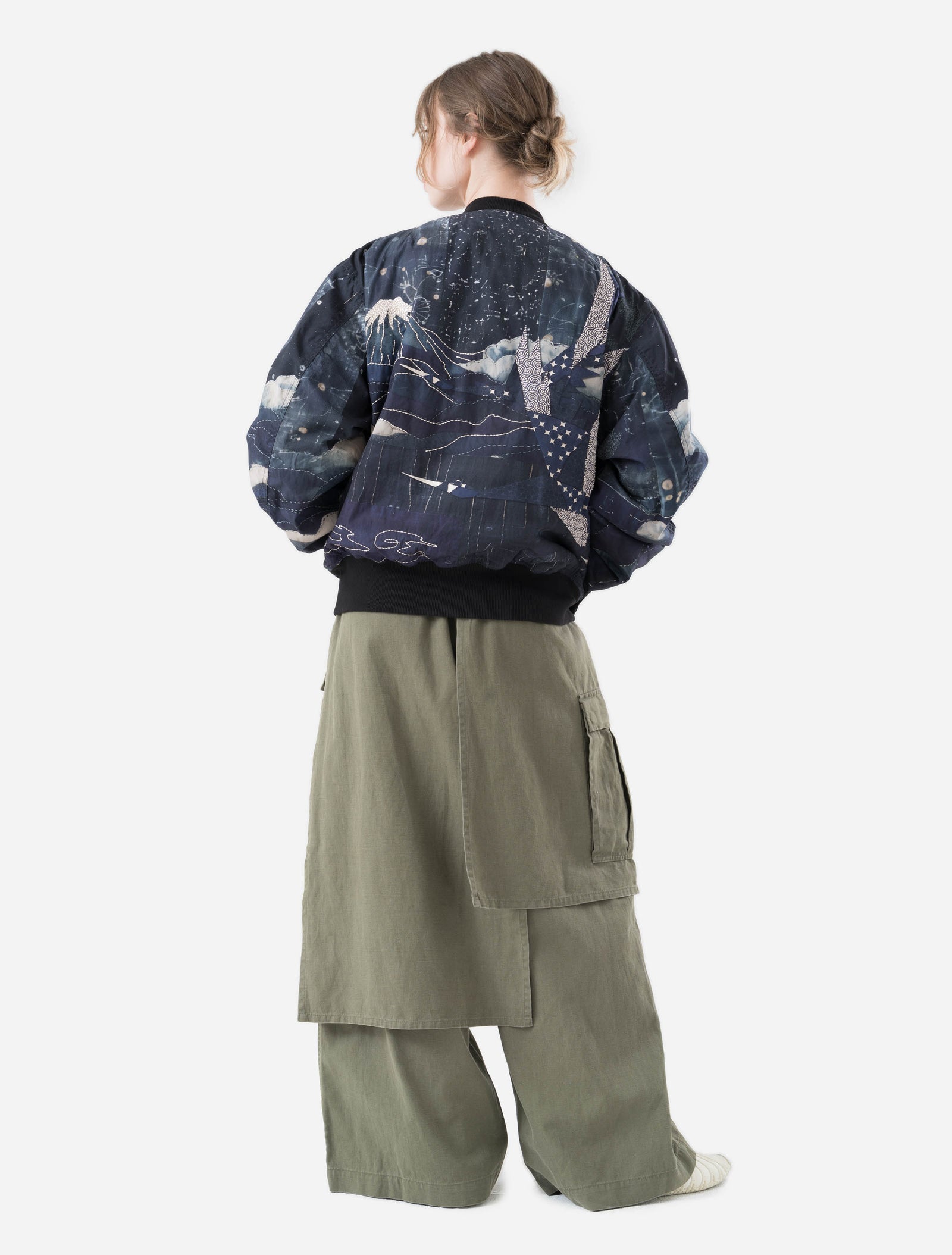 Person wearing a dark blue maharishi embroidered jacket with abstract patterns and light green pants on a white background