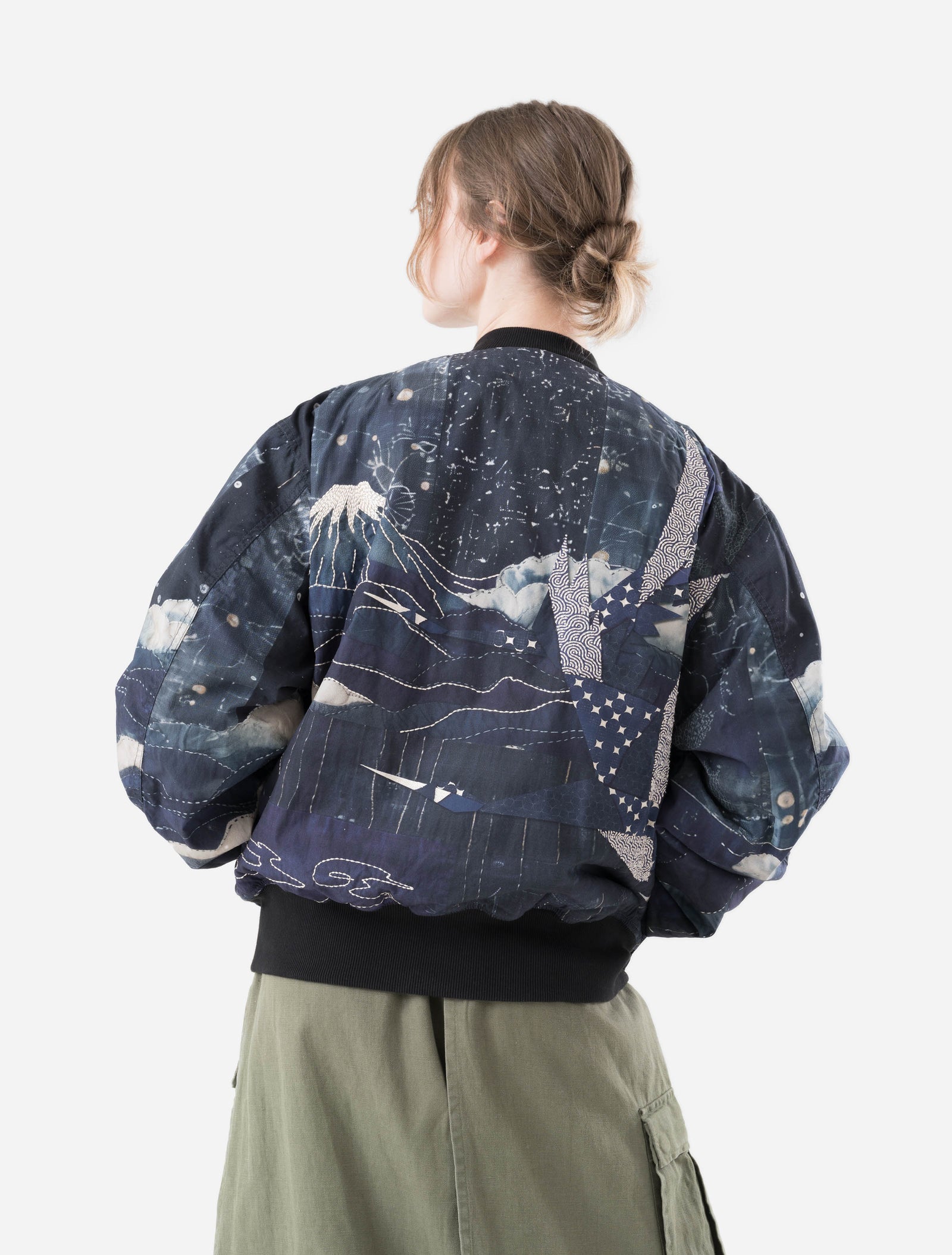 Person wearing a dark blue maharishi embroidered jacket with abstract patterns and light green pants on a white background
