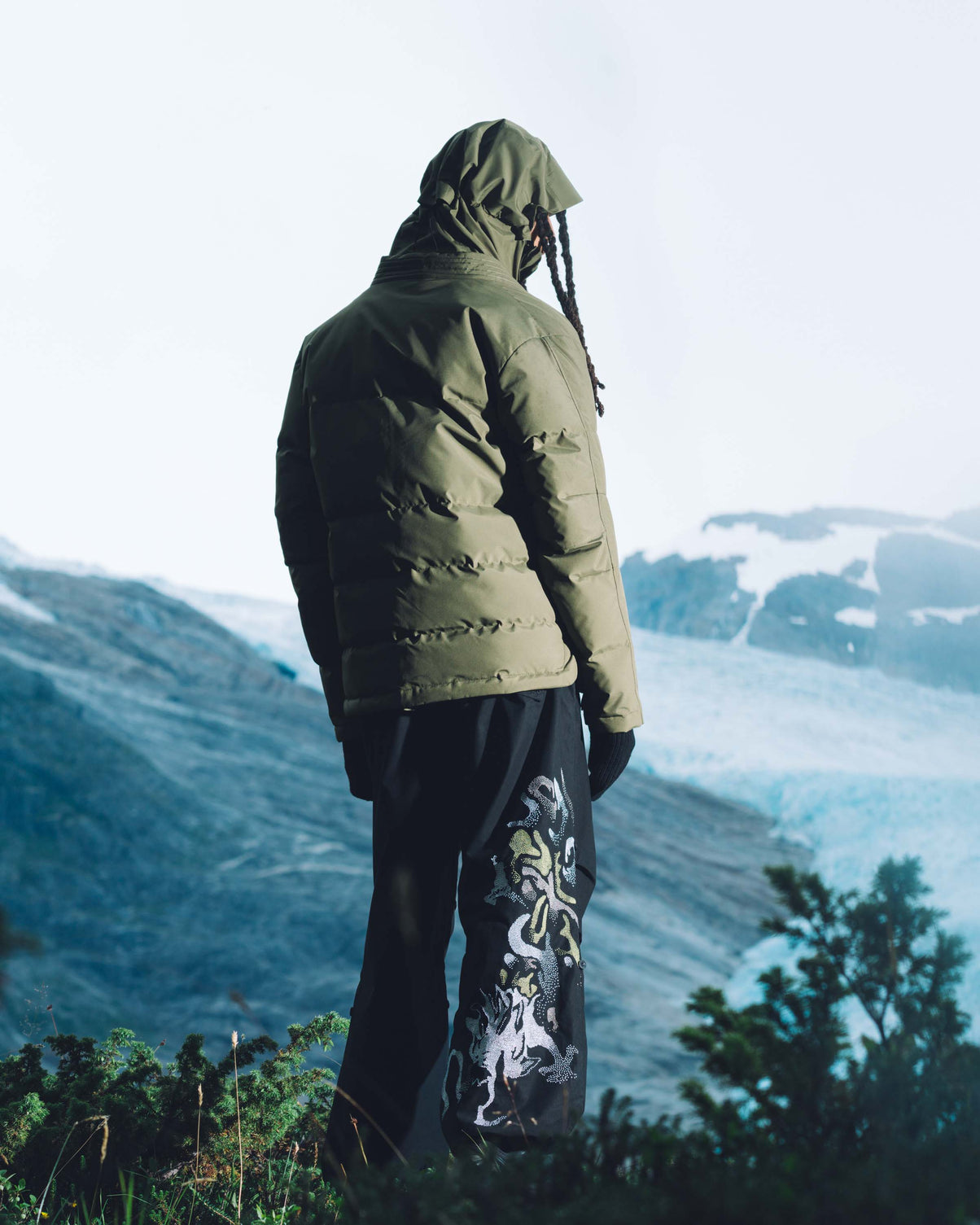 Person wearing a green puffer jacket and black pants with a white design, standing in a mountainous landscape.