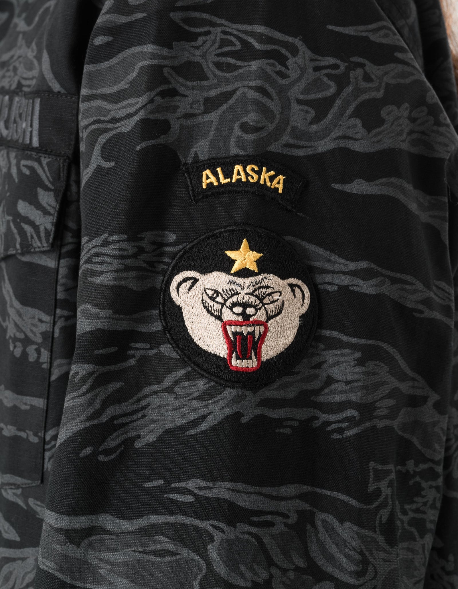 Camouflage jacket with an Alaska patch featuring a bear design.