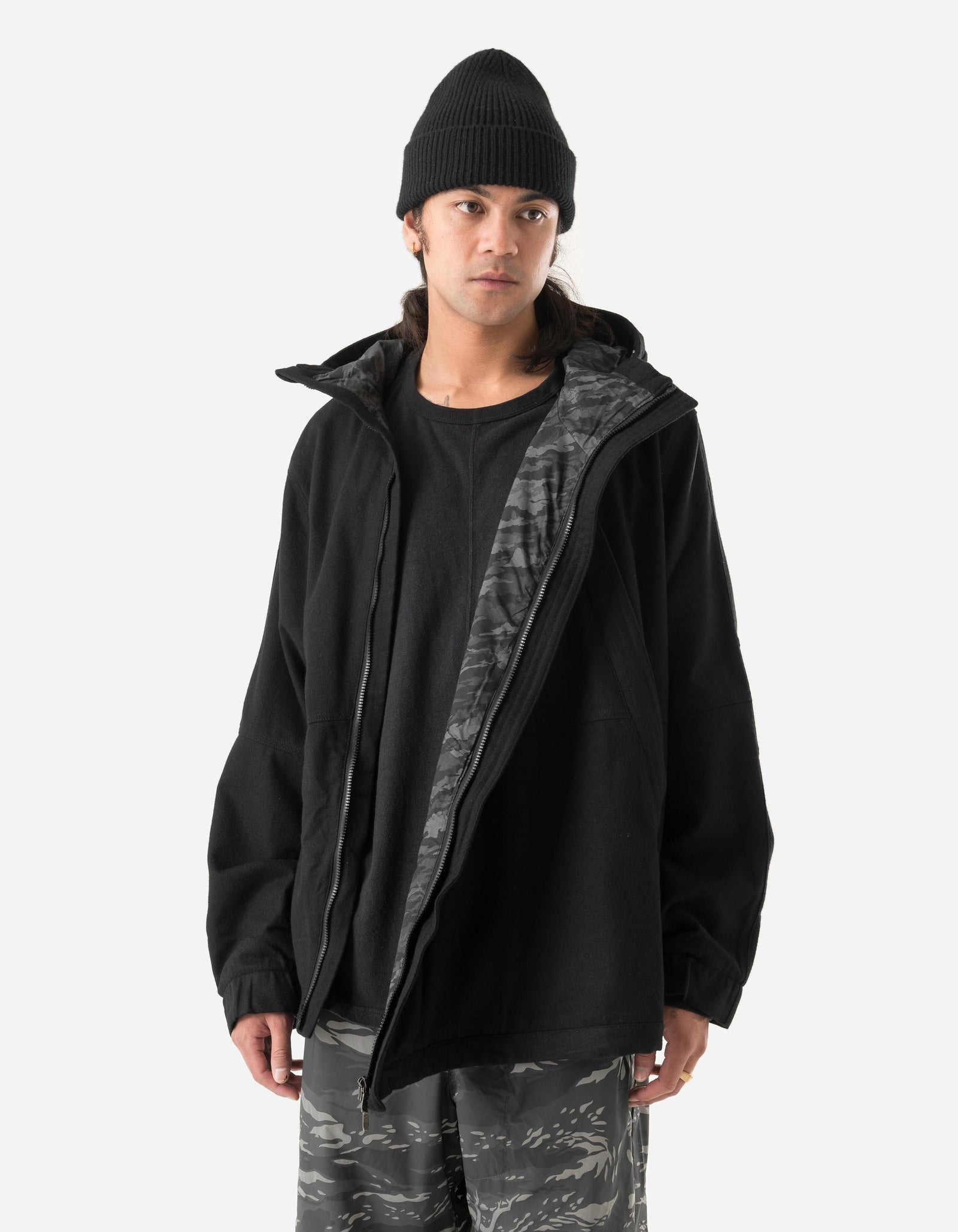 Person wearing a black jacket with a camouflage interior, black beanie, and camouflage pants on a white background