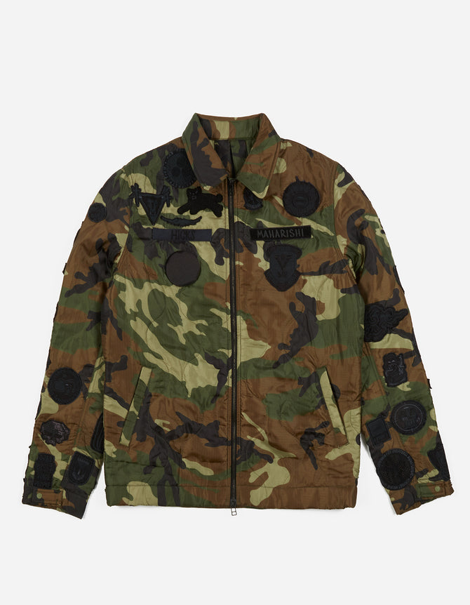 A camouflage quilted liner jacket with blacked out patches and a Maharishi camo pattern. It features a collar, multiple pockets, and a front zipper.